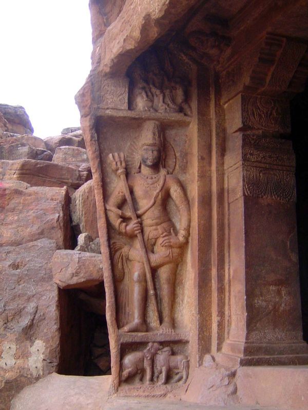A guardian figure or dvarapala. He holds a trident or trisula. Under him, a bull fights an elephant; above him, there's a naga deity. The mo