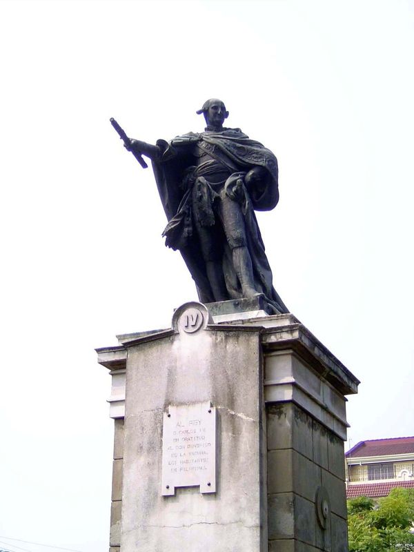 In the center of the square, holding forth to nobody, is Don Carlos IV. The imperial pose is just that, and no more. Charles came to the Spa